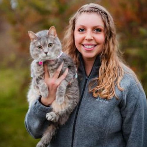 A smiling woman with wavy blonde hair, wearing a gray fleece jacket, holds a gray tabby cat outdoors. As a caring veterinarian, she stands among blurred trees and greenery in the background.