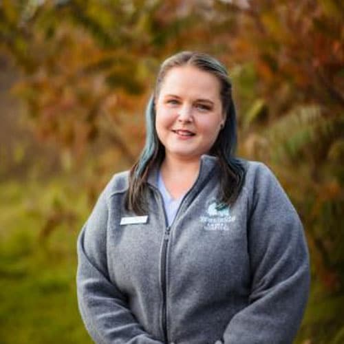 A woman with long brown hair stands outdoors, wearing a gray zip-up jacket with a veterinarian name tag. The background is blurred greenery and autumn-colored foliage. She is smiling gently at the camera.