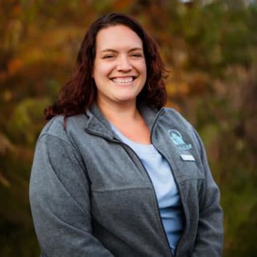 A woman with wavy brown hair is smiling outdoors. She is a veterinarian wearing a gray jacket over a light blue shirt, standing in front of blurred trees with autumn-colored leaves.