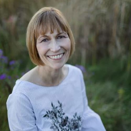 A woman with short light brown hair and a white top with a floral pattern, who is a dedicated veterinarian, smiles while sitting outdoors in a grassy area.