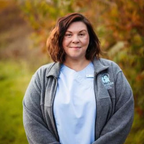 A woman with shoulder-length brown hair smiles at the camera. She is a veterinarian, wearing a light blue shirt and gray jacket. The background is outdoors with greenery and autumn-colored foliage.