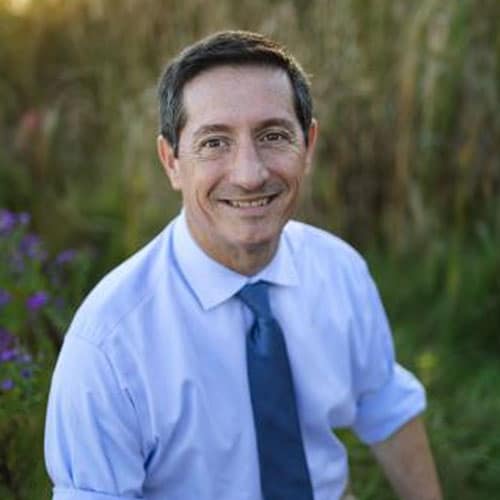 A veterinarian with short dark hair, wearing a light blue shirt and blue tie, smiles while sitting outdoors in front of tall grass and greenery.